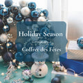 A festive holiday scene with blue and silver Christmas ornaments, a snowman decoration, wrapped gift boxes, string lights, and a large translucent blue label reading 'Holiday Season' and 'Coffret des Fêtes'.